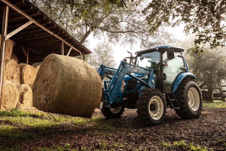 LS Traktor mit Kabine und Frontlader hebt einen Rundballen vor einer Scheune